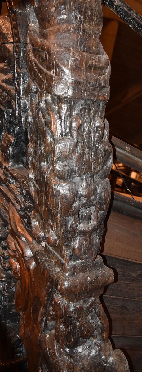 Skulptur i form av en hög, rikt snidad pelare eller stolpe, med kvadratisk genomskärning.

Tre av pelarens sidor är skulpterade medan den fjärde utgör anliggningsyta, försedd med ett antal olikformade urtag. De skulpterade sidorna är horisontellt indelade i tre eller fyra sektioner eller nivåer, avgränsade från varandra av profilerade och snedställda vulster. Nedtill avslutas skulpturen av en ornerad och på mitten insvängd formation.



Ovanför denna nedre avslutning är pelaren utformad som en reslig mansfigur, vänd mot vänster, sedd från betraktaren. Mansfiguren, som täcker tre av skulpturens fyra sidor, är iförd en romersk rustning med en bred fältbindel draperad från höger axel snett över bröstet. På huvudet med det lockiga och utslagna håret sitter en krona av öppen typ, med liljeliknande ornament. Figurens vänstra arm är böjd, medan den högra armen är sträckt utmed sidan. Handen till den högra armen saknas, men har hållit en rikt profilerad sköld, med mycket slitna och vaga konturer av en på bakbenen mot vänster gående grip med stora vingar. Skölden är snidad i ett med figuren, men med kraftiga underskärningar. Vid figurens fötter skymtar ett djur av något slag. Bakom mannens huvud tycks ett snidat textilstycke, kanske del till ett draperi, vara anbragt.



Ovanför den romerska krigaren eller riddaren har pelaren formen av en herm utan armar med långt, krusigt hår och mustaschprytt ansikte. Liksom mansfiguren täcker även hermen tre sidor av pelaren. Huvudet är vridet mot vänster. Överkroppen är bar så när som på ett flikigt plagg som täcker axlar och övre delen av bröstet. Från huvudet och runt midjan är ett tygstycke draperat. Hermens nederdel är formad till en stor maskaron med uppspärrad mun och utsträckt tunga. Maskaronen övergår upptill och utmed sidorna i yppiga bladformationer.



Pelaren avslutas upptill av ett knoppliknande ornament. Ornamentet är ornerat med akantusbladliknande bildningar samt fyra byglar som möts på toppen, där vissa spår antyder att det ursprungligen suttit ett klot eller dylikt, men som fallit bort. Eventuellt kan ornamentet utgöra en huvudbonad till hermen.

Skulpturen är kraftigt sliten.



Text in English: Sculpture in the shape of a richly carved column or pillar.

Three sides of the pillar are carved, while the fourth side comprises the attachment surface and has a number of variously-shaped indentations. The sculptured sides are divided horizontally into three or four sections separated by angled tori.



The lower section of the pillar is carved in the figure of a tall man. The figure is clad in Roman armour and has a field badge (cloak/plaid) draped across his chest at an angle from his right shoulder. On his head he wears an open crown. He has long, curly hair. The right arm is broken off, the left is at his side. The left hand is missing but once held a richly carved shield.

The shield is carved out of the same piece of wood as the figure, but deeply undercut. A carved figure is apparent on the shield. Although the shield is badly worn, it is possible to recognise one of the hind legs of a griffin with large wings. At the man's feet lies an animal of some kind.



Above the Roman warrior or knight, the pillar is carved in the shape of a herm, without arms and with long, curly hair and a moustache. Like the male figure, the herm also covers three sides of the pillar. The head is twisted to the left. The upper part of the body is bare except for a piece of cloth that covers the shoulders and upper chest. The lower part of the herm is carved as a large mask, its mouth open and tongue protruding. The sides and upper parts of the mask are carved as luxuriant leaves.



The upper part of the pillar has a ornament in shape of a knob. The ornament has a ring of acanthus leaves and four spherical loops run vertically from the acanthus leaves and meet at a point at the top. An object, for example a globe appears to have adorned the top of the ornament. This object is missing. The ornament may also be a part of the headgear of the herm.

The sculpture is severely worn.