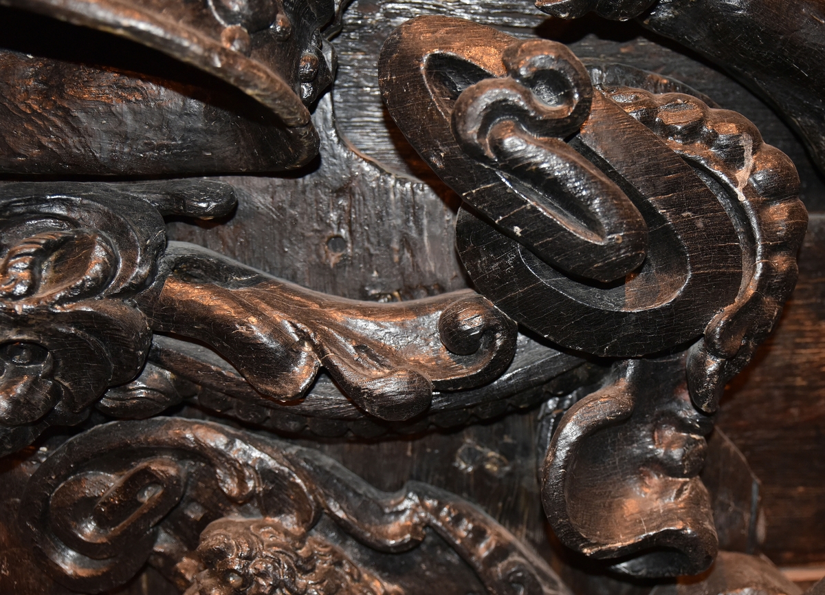 Broskornament. Ornamentet är utformat som ett kattdjursliknande huvud från vilket utgår "armar", dekorerade med blad och broskvoluter. Ändpartierna är förtjockade med elipsformade broskvoluter, blad och nedhängande flikar. Baksidan är slät.
Ornamentet är brutet på baksidans mittparti.

Text in English: Earshell ornament consisting of a cat-like head, from which extend "arms" decorated with ellipse-shaped pearlwork scroll leaves and downward-hanging leaf-blades with thickened ends. The reverse side is smooth.
The ornament is broken in the middle on the reverse side.