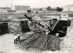 Omkring mitten av 1940-talet började allmänt grävmaskiner användas vid schakt av husgrunder. Grävmaskinen på bilden utför schakt till panncentralen i kvarteret Lien, Wettterlinsgatan 19. Panncentralen betjänar 260 lägenheter i kvarteren Plogen, Harven, Lien och Räfsan med värme och varmvatten. Under ett tunnare lager av matjord var det 3,5 meter djup pinnmo ned till kalk-berget. Ytan ovanpå kalkberget var i allmänhet plan med horisontala och vertikala sprickor. Grävmaskinen lösgjorde och lastade jorden på lastbilar. Två män följde grävmaskinen och skyfflade fram den jord som maskinen inte kunde få med i skopan. Kalkberget borrades sedan och sprängdes ytterligare 3 meter djupare, varefter kalkstensskrotet med grävmaskinen lastades på lastbilar. En del av detta material användes som underlag till vägar och gator. Till vänster på bilden en halv meter under markytan är brädor synliga. De utgjorde skydd över rikskabel från Stockholm och söderut.