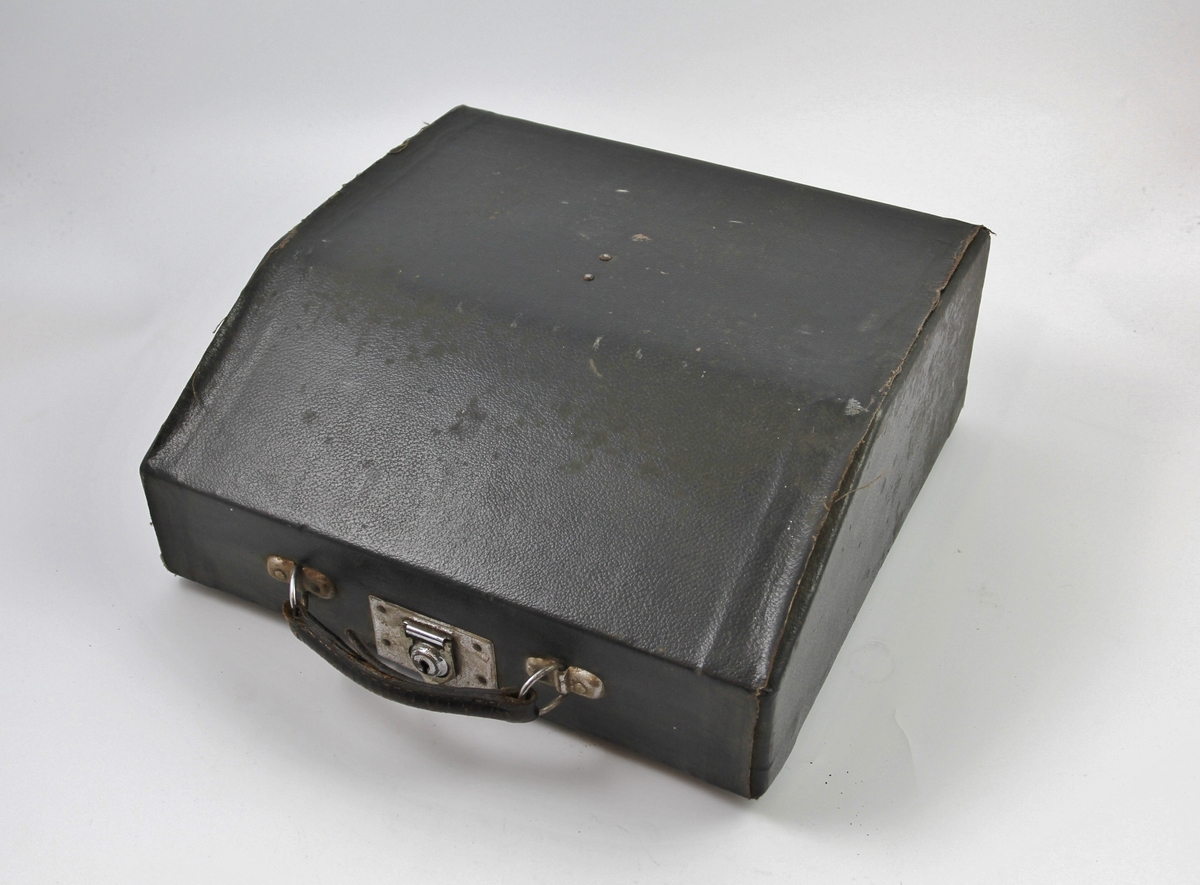 Skrivemaskin med beskyttelseshette. Norske bokstaver med litt annen bunnfarge enn de øvrige bokstavene, tyske termer på funksjonsknapper. Svartlakkert metall, lyse knapper. Beskyttelseshette i svart skinn med håndtak og smekklås.