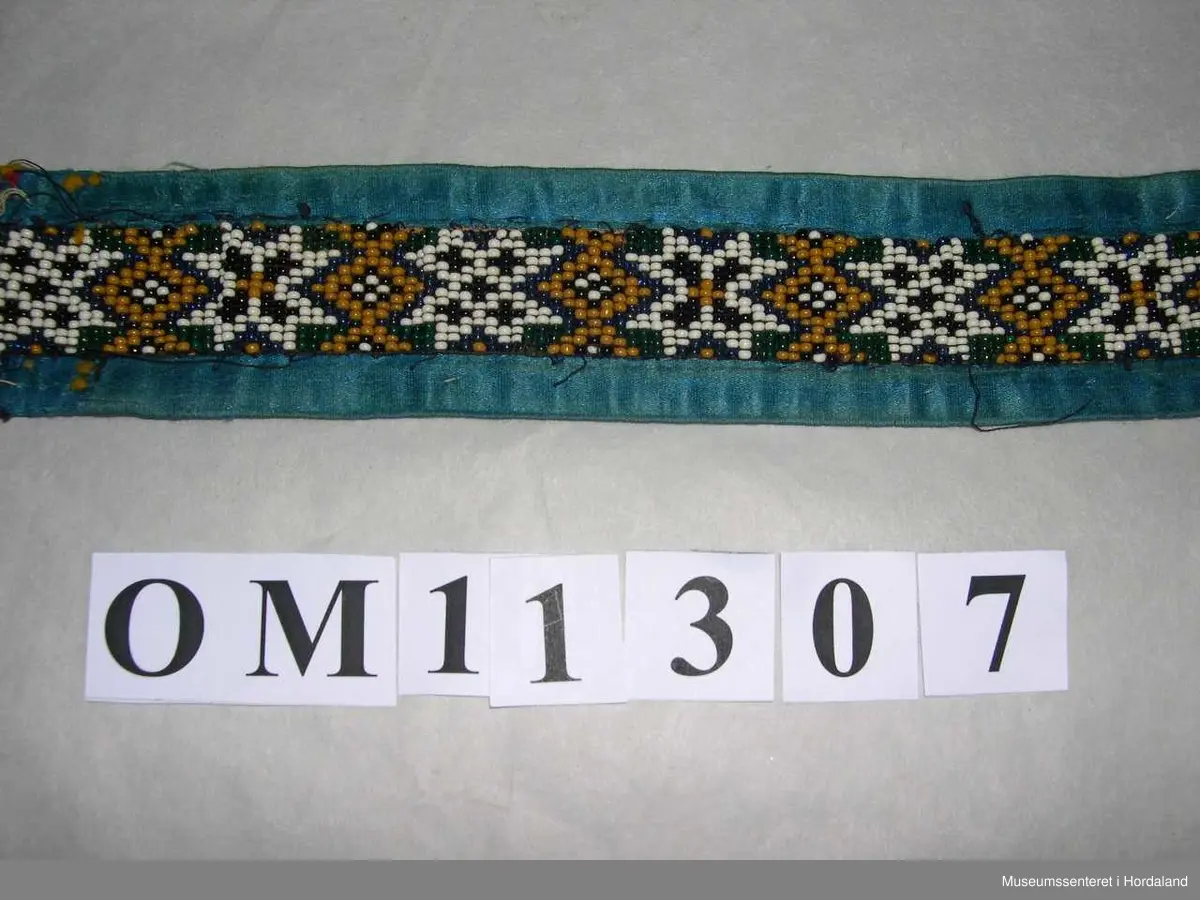 Perlebelte vove på perlegogn og montert på eit brunrosa bakstykke med blåe silkeband rundt kanten. Perledelen går litt utom kantbandet i eine enden. Motivet på perleveven er åttebladroser.