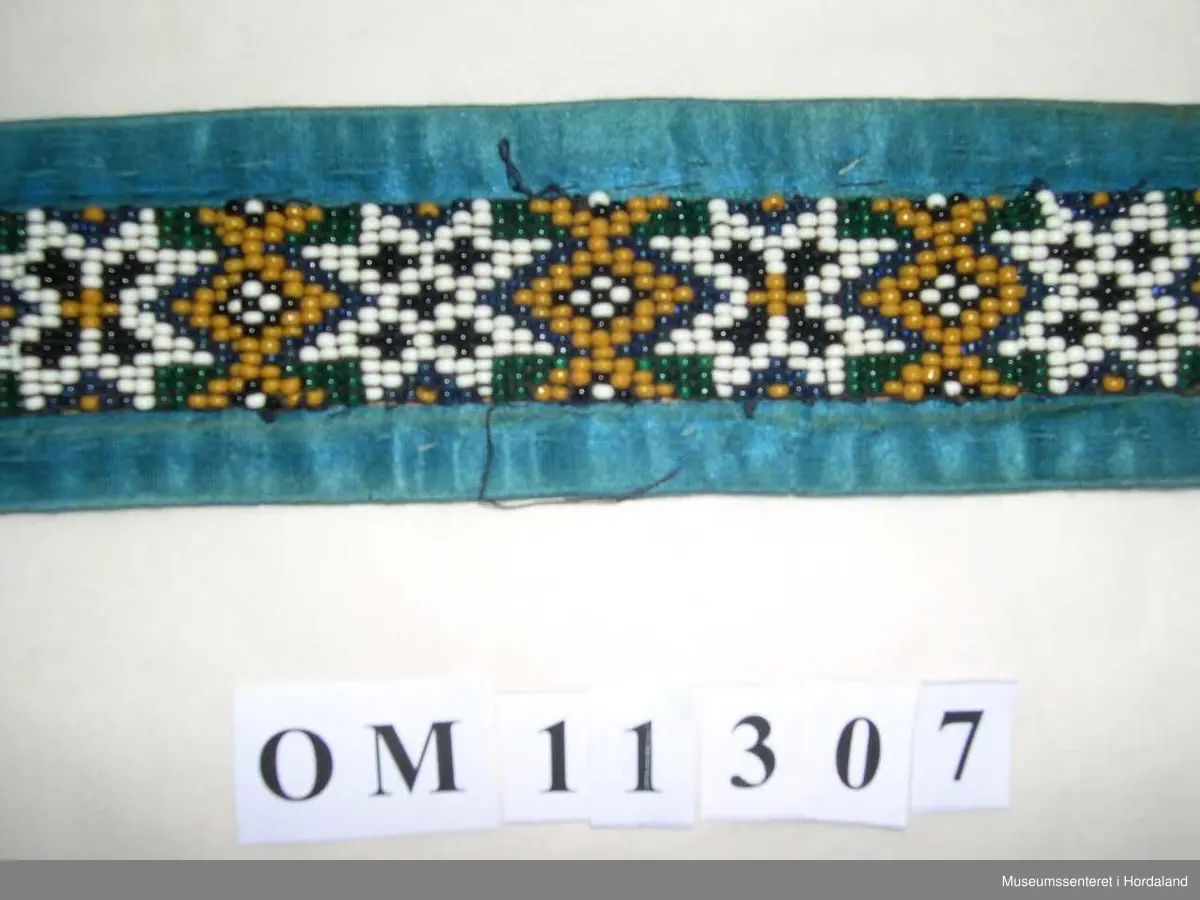 Perlebelte vove på perlegogn og montert på eit brunrosa bakstykke med blåe silkeband rundt kanten. Perledelen går litt utom kantbandet i eine enden. Motivet på perleveven er åttebladroser.