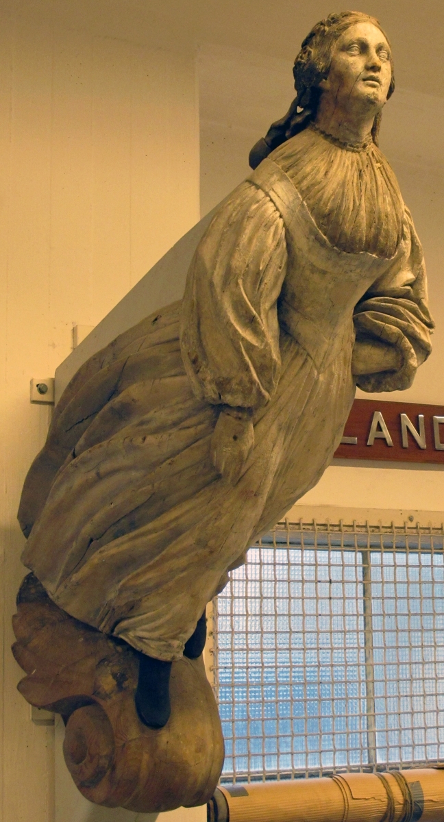 Galjonsbild, kvinna i helfigur med vid kjol i svepande veck, gammal vit kridering. Mörkt hår i hårnät samt svart fotbeklädnad.