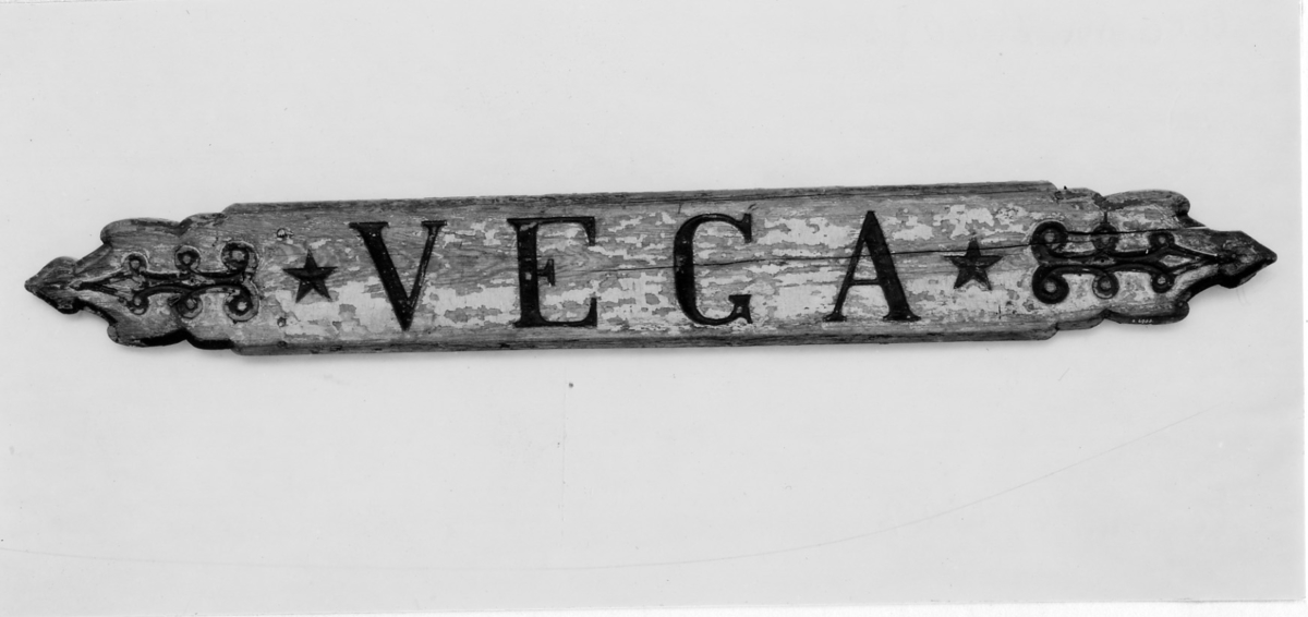Namnbräda, "VEGA", inskrivet (rosartversaler) + femuddiga stjärnor, pilliknande dekor. Fasad kant, skulpterad i ändstyckena. Starkt avflagnad, grå färg. Text och dekor i svart.