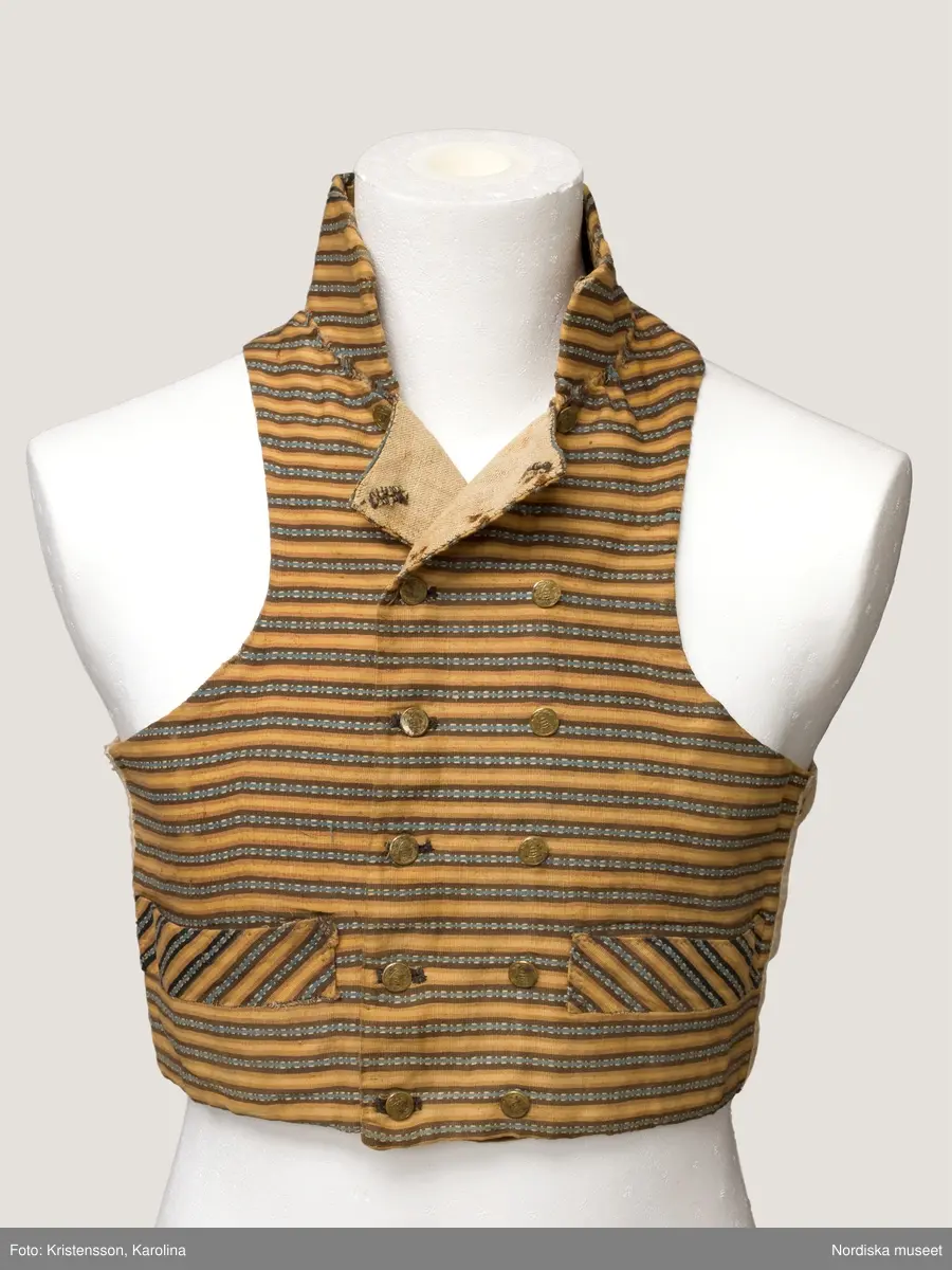 Väst i kort modell av randig bomullsrips i brunt och beige med invävd mönsterrand i den mörkbruna randen i ljusblått med vita "hoppor". Västen är tvärrandig med 2 framstycken och hög styvad  krage av samma tyg, ryggstycke av fin oblekt linnelärft, foder av grov och gles linnelärft i ryggen och finare i framstyckena, 2 stolpfickor, dubbelknäppning med knapphål på båda framstyckena och 6 par mässingsknappar med Ångermanlands landskapsvapen med 3 liggande laxar.
Tygsorten finns under benämningen: Västtyg i flera av 1820-30 talets tygprovsböcker och har enligt uppgift använts som brudgumsväst 1824
/Berit Eldvik  2005-11