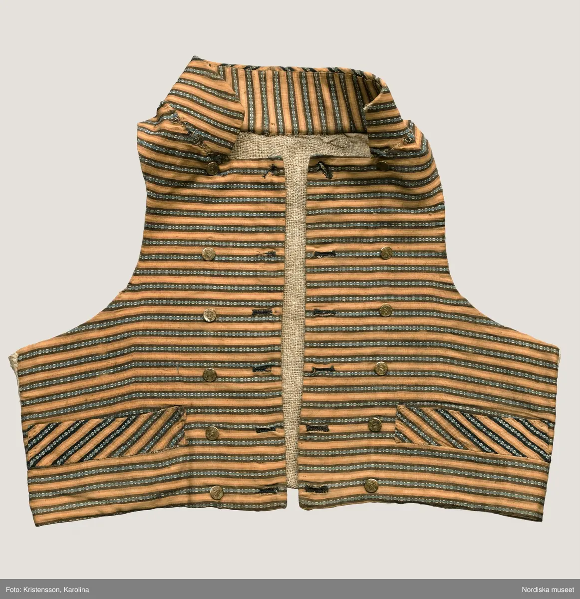 Väst i kort modell av randig bomullsrips i brunt och beige med invävd mönsterrand i den mörkbruna randen i ljusblått med vita "hoppor". Västen är tvärrandig med 2 framstycken och hög styvad  krage av samma tyg, ryggstycke av fin oblekt linnelärft, foder av grov och gles linnelärft i ryggen och finare i framstyckena, 2 stolpfickor, dubbelknäppning med knapphål på båda framstyckena och 6 par mässingsknappar med Ångermanlands landskapsvapen med 3 liggande laxar.
Tygsorten finns under benämningen: Västtyg i flera av 1820-30 talets tygprovsböcker och har enligt uppgift använts som brudgumsväst 1824
/Berit Eldvik  2005-11