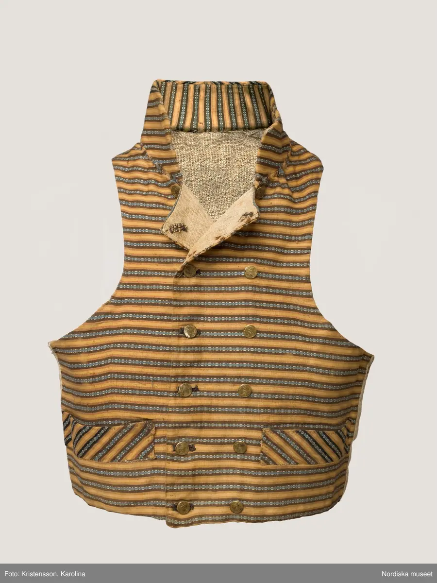 Väst i kort modell av randig bomullsrips i brunt och beige med invävd mönsterrand i den mörkbruna randen i ljusblått med vita "hoppor". Västen är tvärrandig med 2 framstycken och hög styvad  krage av samma tyg, ryggstycke av fin oblekt linnelärft, foder av grov och gles linnelärft i ryggen och finare i framstyckena, 2 stolpfickor, dubbelknäppning med knapphål på båda framstyckena och 6 par mässingsknappar med Ångermanlands landskapsvapen med 3 liggande laxar.
Tygsorten finns under benämningen: Västtyg i flera av 1820-30 talets tygprovsböcker och har enligt uppgift använts som brudgumsväst 1824
/Berit Eldvik  2005-11