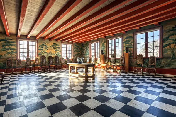 Foto av barokkt interiør på Linderud gård. Sjakkmønstret gulv, veggmalerier og røde takbjelker. I rommet står et trebord med en urne. 