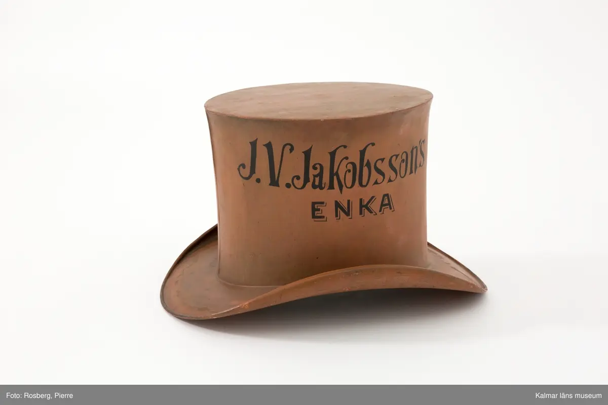 KLM 6304. Hattmakareskylt. Stor rödbrun hatt (s. k. storm) av järnbleck med svart text målad på båda sidor: J.V. Jakobssons ENKA. Tillhörande enkel järnarm för fäste på vägg. Armen är grönlackerad och tillverkad senare, omkring 1995.
