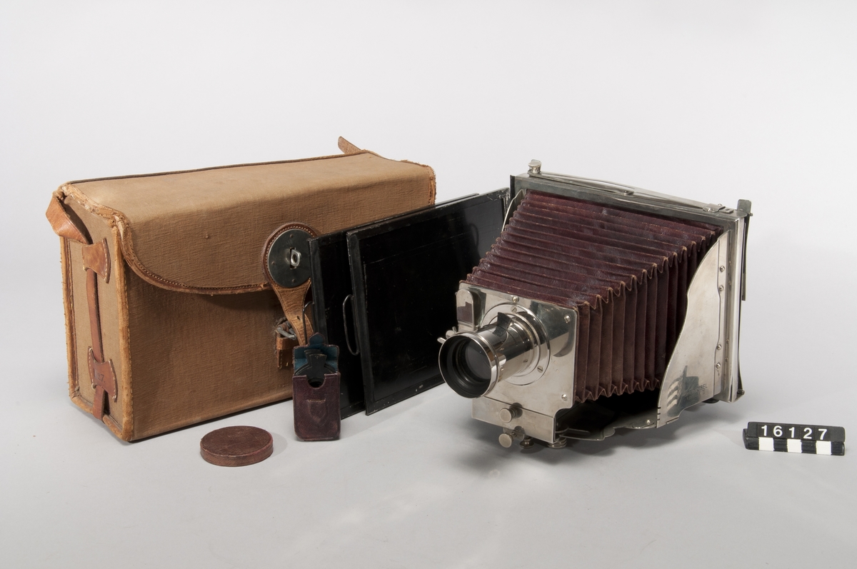 This was the first metal folding camera, the Model II. It was designed with a patent by H. Mader in 1888-1890. The camera was donated to the museum in January 1939 by Helmer Bäckström, who was a professor of photography. The camera is marked "5" in the slats of the bellows as well as with a round mark with the letters G M R and two sword-like characters. The lens is marked "Aplanal Invincible" and has "VI" engraved on the back at the threads. The camera comes with four cassettes, five plug-in apertures and a textile-clad case.