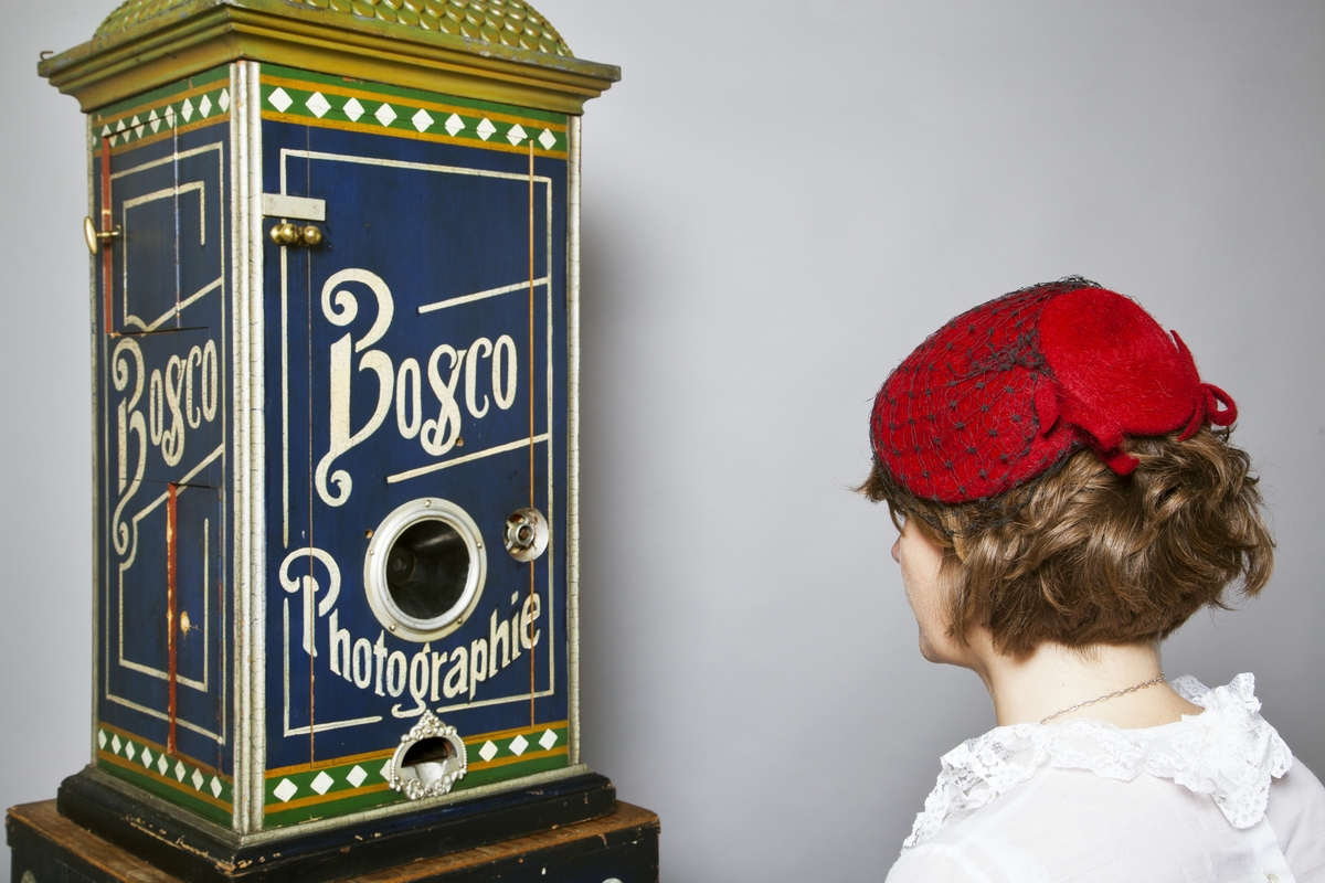 Fotoautomat "Bosco" för tillverkning av ferrotypier. Penningautomat för självfotografering.  Den fungerade på så sätt att det ljuskänsliga skiktet placerats i en profilerad plåtbricka som på samma gång var framkallningsskål och sedan ram för fotografiet. I automaten exponerades bilden i lodrätt läge och framkallades sedan med vätskor som portionerades ur behållare. Efter tre minuter var ferrotypen klar och matades ut ur apparaten. Den behövde torka ytterligare och fick enligt anvisningarna inte utsättas för starkt ljus förrän den var torr. Bilden mätte 60x83 mm och levererades i ett etui av papper. Plåtbrickan hade påtryck av reklam och information på baksida och kanter.
Tillbehör: Sockel med 2 st. metallådor för plåtar, 10 st. plåtar ("W.H.C Bernitt Original Bosco Platte Hamburg 23 ...), överdel med kamera, mekanism, mekanism för framkallning m.m. nyckel till verket.