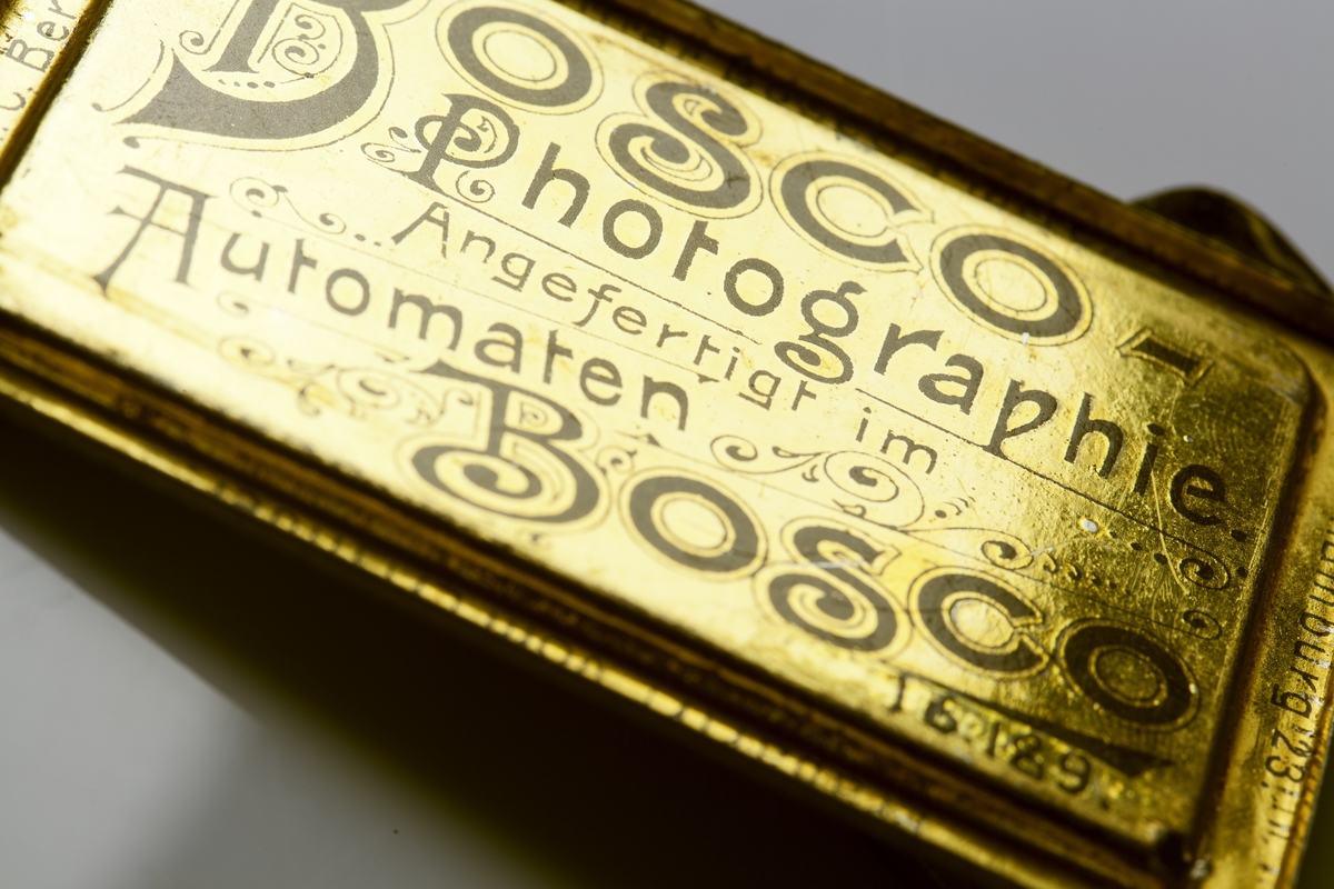 Fotoautomat "Bosco" för tillverkning av ferrotypier. Penningautomat för självfotografering.  Den fungerade på så sätt att det ljuskänsliga skiktet placerats i en profilerad plåtbricka som på samma gång var framkallningsskål och sedan ram för fotografiet. I automaten exponerades bilden i lodrätt läge och framkallades sedan med vätskor som portionerades ur behållare. Efter tre minuter var ferrotypen klar och matades ut ur apparaten. Den behövde torka ytterligare och fick enligt anvisningarna inte utsättas för starkt ljus förrän den var torr. Bilden mätte 60x83 mm och levererades i ett etui av papper. Plåtbrickan hade påtryck av reklam och information på baksida och kanter.
Tillbehör: Sockel med 2 st. metallådor för plåtar, 10 st. plåtar ("W.H.C Bernitt Original Bosco Platte Hamburg 23 ...), överdel med kamera, mekanism, mekanism för framkallning m.m. nyckel till verket.