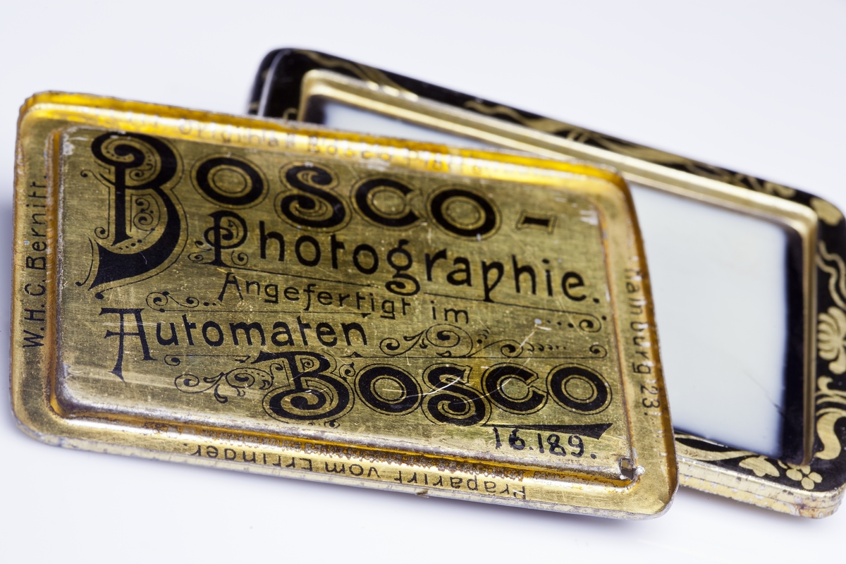 Fotoautomat "Bosco" för tillverkning av ferrotypier. Penningautomat för självfotografering.  Den fungerade på så sätt att det ljuskänsliga skiktet placerats i en profilerad plåtbricka som på samma gång var framkallningsskål och sedan ram för fotografiet. I automaten exponerades bilden i lodrätt läge och framkallades sedan med vätskor som portionerades ur behållare. Efter tre minuter var ferrotypen klar och matades ut ur apparaten. Den behövde torka ytterligare och fick enligt anvisningarna inte utsättas för starkt ljus förrän den var torr. Bilden mätte 60x83 mm och levererades i ett etui av papper. Plåtbrickan hade påtryck av reklam och information på baksida och kanter.
Tillbehör: Sockel med 2 st. metallådor för plåtar, 10 st. plåtar ("W.H.C Bernitt Original Bosco Platte Hamburg 23 ...), överdel med kamera, mekanism, mekanism för framkallning m.m. nyckel till verket.