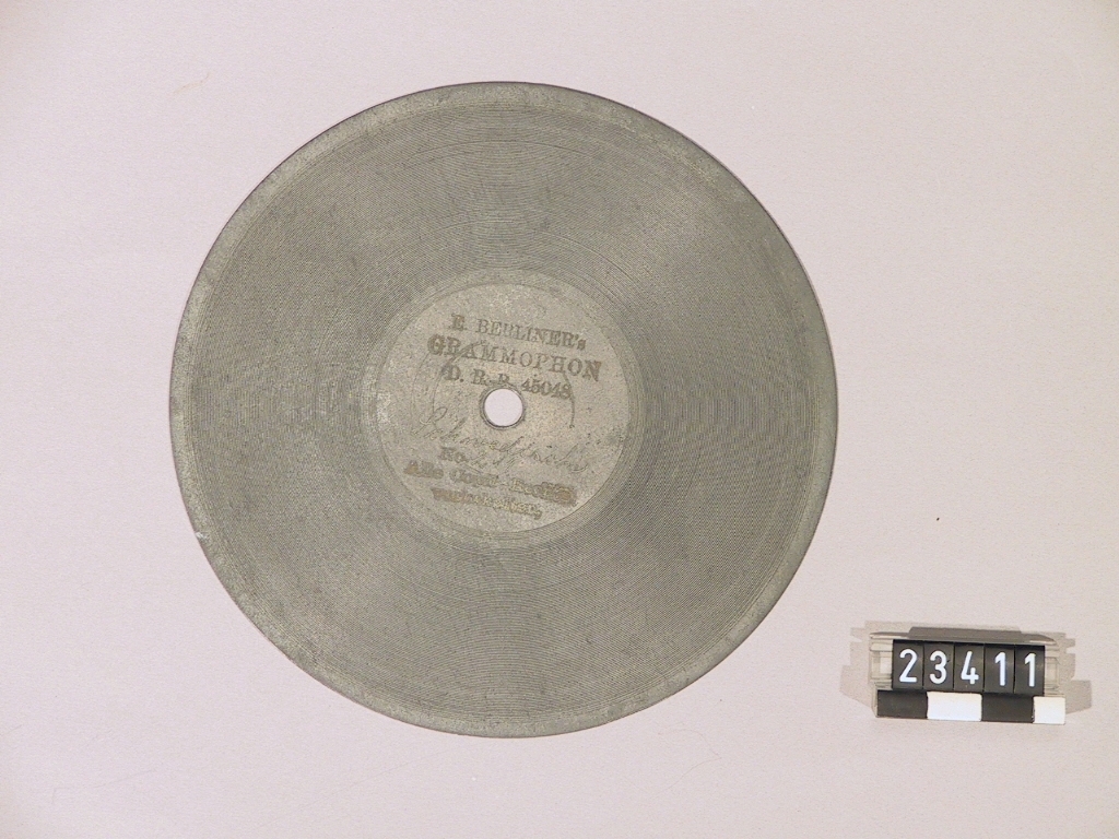 Grammofon av den första kommersiella typen. Bottenplattan märkt: DRP 45048. Tratt av mässing, stämplad: "1778". Trattmynningen stämplad med nummer "62". På insidan är tratten målad med röd färg. Trattens främre del skall vara riktad snett uppåt genom att den ligger på ett stöd som på detta exemplar saknas. Ingår även 1st. skiva