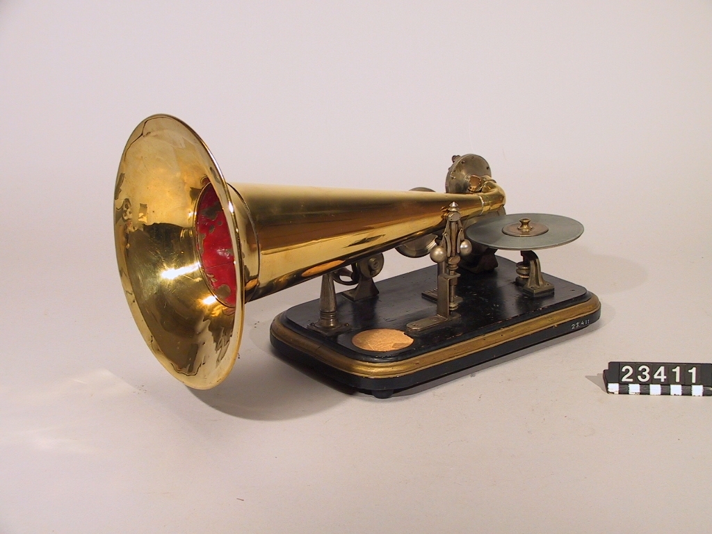 Grammofon av den första kommersiella typen. Bottenplattan märkt: DRP 45048. Tratt av mässing, stämplad: "1778". Trattmynningen stämplad med nummer "62". På insidan är tratten målad med röd färg. Trattens främre del skall vara riktad snett uppåt genom att den ligger på ett stöd som på detta exemplar saknas. Ingår även 1st. skiva