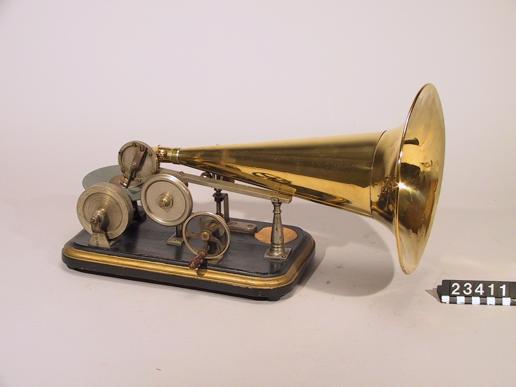 Grammofon av den första kommersiella typen. Bottenplattan märkt: DRP 45048. Tratt av mässing, stämplad: "1778". Trattmynningen stämplad med nummer "62". På insidan är tratten målad med röd färg. Trattens främre del skall vara riktad snett uppåt genom att den ligger på ett stöd som på detta exemplar saknas. Ingår även 1st. skiva
