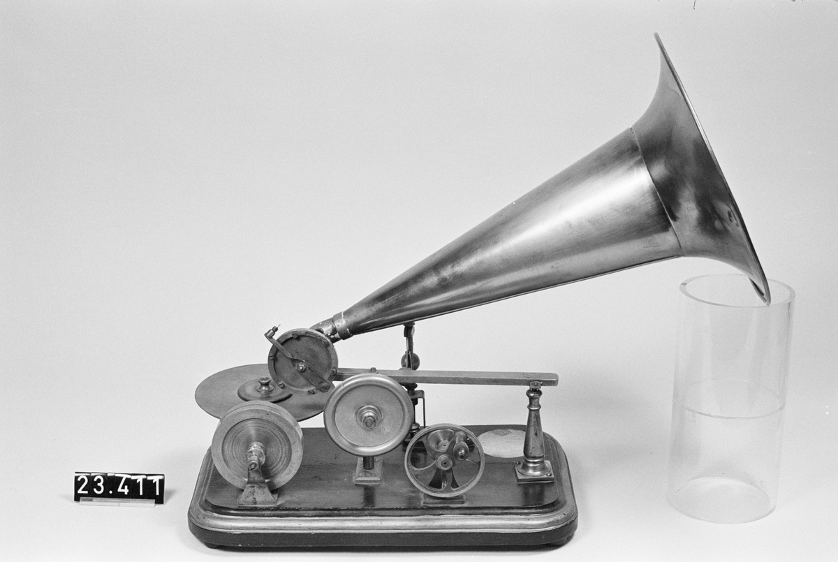 Grammofon av den första kommersiella typen. Bottenplattan märkt: DRP 45048. Tratt av mässing, stämplad: "1778". Trattmynningen stämplad med nummer "62". På insidan är tratten målad med röd färg. Trattens främre del skall vara riktad snett uppåt genom att den ligger på ett stöd som på detta exemplar saknas. Ingår även 1st. skiva