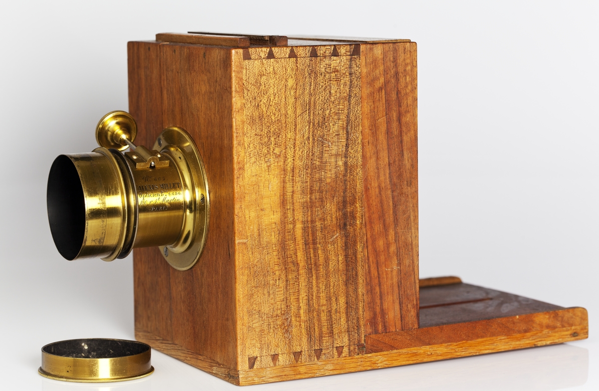 Kamera, skjutlådemodell. Våtplåt 9,5 x 11,5. Objektiv: Alexis Millet, Opticien Bte'SGDG 160r.st:Martin Paris. Nr 605. Utrustning för fotografering på resor, jämte koffert. Lösa delar: kamera av trä med objektiv från Alexis Millet, Paris, nr. 605, objektivlock av mässing, visirskiva, 4 kassetter för våtplåtar 9 x 12 cm, bakstycke med visirskiva för stereobilder 8 x 8 cm, 2 kassetter för stereovåtplåtar, kärl av zinkplåt med lock (gångjärnet avbrutet), trattformat kärl av zinkplåt, mindre kuvert av glas (spräckt och lagad, troligen av givaren), större kuvert av glas, torkställ av trä, 2 skivor av trä med spår för plåtar, kopieram för max 16 x 20 cm, vikbar skiva av trä (för beredning, troligen av våtplåtar), liten bordskiva med kantlist (passar till stativet TM 25.111,fodral av trä med 12 st. våtplåtnegativ 9 x 12 cm (porträtt och skulpturer), fodral av trä med 12 st. sämre negativ, fodral av trä med 10 st. blanka och  skrapade glasplåtar, diverse plåtar i paket (i ask ny 1950), fodral av trä med 11 st. stereovåtplåtar, varav 3 preparerade.
Tillbehör: Koffert.