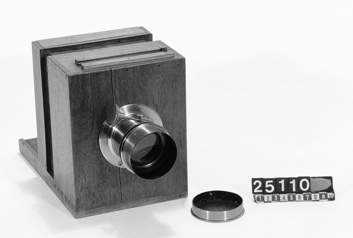 Kamera, skjutlådemodell. Våtplåt 9,5 x 11,5. Objektiv: Alexis Millet, Opticien Bte'SGDG 160r.st:Martin Paris. Nr 605. Utrustning för fotografering på resor, jämte koffert. Lösa delar: kamera av trä med objektiv från Alexis Millet, Paris, nr. 605, objektivlock av mässing, visirskiva, 4 kassetter för våtplåtar 9 x 12 cm, bakstycke med visirskiva för stereobilder 8 x 8 cm, 2 kassetter för stereovåtplåtar, kärl av zinkplåt med lock (gångjärnet avbrutet), trattformat kärl av zinkplåt, mindre kuvert av glas (spräckt och lagad, troligen av givaren), större kuvert av glas, torkställ av trä, 2 skivor av trä med spår för plåtar, kopieram för max 16 x 20 cm, vikbar skiva av trä (för beredning, troligen av våtplåtar), liten bordskiva med kantlist (passar till stativet TM 25.111,fodral av trä med 12 st. våtplåtnegativ 9 x 12 cm (porträtt och skulpturer), fodral av trä med 12 st. sämre negativ, fodral av trä med 10 st. blanka och  skrapade glasplåtar, diverse plåtar i paket (i ask ny 1950), fodral av trä med 11 st. stereovåtplåtar, varav 3 preparerade.
Tillbehör: Koffert.