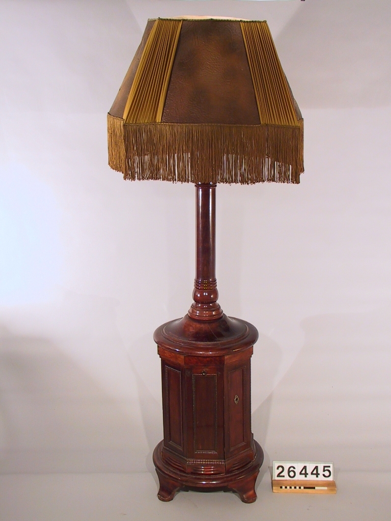 Stor golvlampa med grammofon. Ljudet leds i piedestalen till ljudöppning under lampskärmen. Åttkantigt underskåp av mahogny och mahognybetsad bok, med inredning av ek, innehållande fjäderdrivet grammofonverk och ljudarm av trä. I ytterväggen finns hål för vev. Ihålig pelare, upptill avslutad med pyramidformad ljudtratt av trä med fyra glödlampor. Däröver en fyrkantig (buktig) lampskärm av mönsterpressat "pergamentpapper" och siden med silkesfrans. Skärmen (spec. sidenet) mycket skadad. Skärmens diameter: ca. 800 mm. Grammonfonen stannar själv då skivan är slutspelad. Nu befintlig elektrisk ljuddosa monterad senare i stället för ursprunglig membrandosa (som saknas).
Tillbehör: Vev.