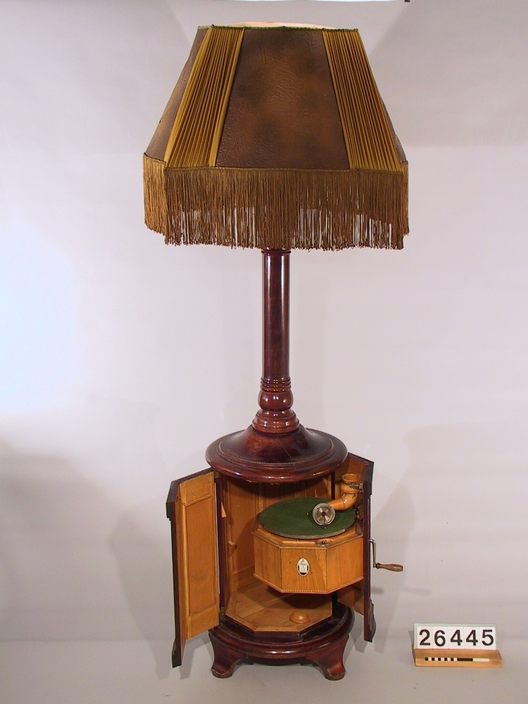 Stor golvlampa med grammofon. Ljudet leds i piedestalen till ljudöppning under lampskärmen. Åttkantigt underskåp av mahogny och mahognybetsad bok, med inredning av ek, innehållande fjäderdrivet grammofonverk och ljudarm av trä. I ytterväggen finns hål för vev. Ihålig pelare, upptill avslutad med pyramidformad ljudtratt av trä med fyra glödlampor. Däröver en fyrkantig (buktig) lampskärm av mönsterpressat "pergamentpapper" och siden med silkesfrans. Skärmen (spec. sidenet) mycket skadad. Skärmens diameter: ca. 800 mm. Grammonfonen stannar själv då skivan är slutspelad. Nu befintlig elektrisk ljuddosa monterad senare i stället för ursprunglig membrandosa (som saknas).
Tillbehör: Vev.