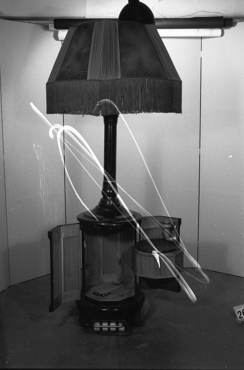Stor golvlampa med grammofon. Ljudet leds i piedestalen till ljudöppning under lampskärmen. Åttkantigt underskåp av mahogny och mahognybetsad bok, med inredning av ek, innehållande fjäderdrivet grammofonverk och ljudarm av trä. I ytterväggen finns hål för vev. Ihålig pelare, upptill avslutad med pyramidformad ljudtratt av trä med fyra glödlampor. Däröver en fyrkantig (buktig) lampskärm av mönsterpressat "pergamentpapper" och siden med silkesfrans. Skärmen (spec. sidenet) mycket skadad. Skärmens diameter: ca. 800 mm. Grammonfonen stannar själv då skivan är slutspelad. Nu befintlig elektrisk ljuddosa monterad senare i stället för ursprunglig membrandosa (som saknas).
Tillbehör: Vev.