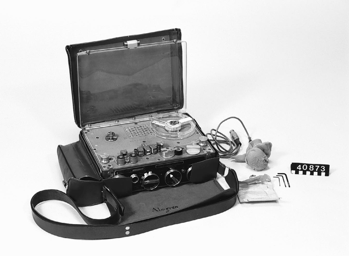 Reportagebandspelare Kudelski Nagra III Pilot för ¼ " magnetband. Ett inspelat band med intervju från 1962 med Algeriets upprorsledare Ben Bella medföljer. Tillhörande mikrofon, läderväska samt tre insexnycklar. Bandspelaren drivs med 9 st 1,5 V batterier.  Bandspelarens pilottonsfunktion gör att den kan kopplas till filmkamera för synkronisering av ljud och bild. Mikrofonen är försedd med ett skumgummiöverdrag för att motverka vindbrus och "puffar" av hårda konsonanter vid intervju.  Metoden för synkronisering uppfanns av Stefan Kudelski. Pilottonen spelas in på två 0,45 mm breda spår med 0,4 mm avstånd i ljudspåret. Genom att signalen är i motfas hörs inte signalen när bandet spelas upp.
Tillbehör: Mikrofon, tomspole, väska, 3 st .insexnycklar, inspelat magnetband