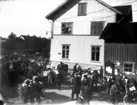 Marknadsdag i Floby centrum. I byggnaden David Karlssons speceri- och diversehandel i den västra, och i den östra järnhandeln som förestods av Teodor Johansson. Mitt i bilden August Johansson i Qvissle.