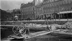 Handdriven grävmaskin i arbete i Malmö hamn. Använd till 1914. Konstruktion från 1770-talet. I bakgrunden skymtar Jernvägs-Hotellet. (Bilden är publicerad i Daedalus 1941).