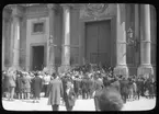 Skioptikonbild från institutionen för fotografi vid Kungliga Tekniska Högskolan. Bilden är troligen tagen av John Hertzberg under en resa i Europa. Hertzberg blev docent i fotografi 1921vid Kungliga Tekniska Högskolan i Stockholm. Folksamling utanför en port.