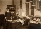 Fotografi ur album tillhörande Ester Holmberg, anställd vid Telegrafstyrelsens statistikavdelning 1902. Fotografierna troligen tagna 1907. Agnes Forsbeck, Bch Hedén och Elin Lidman vid skrivbordet.