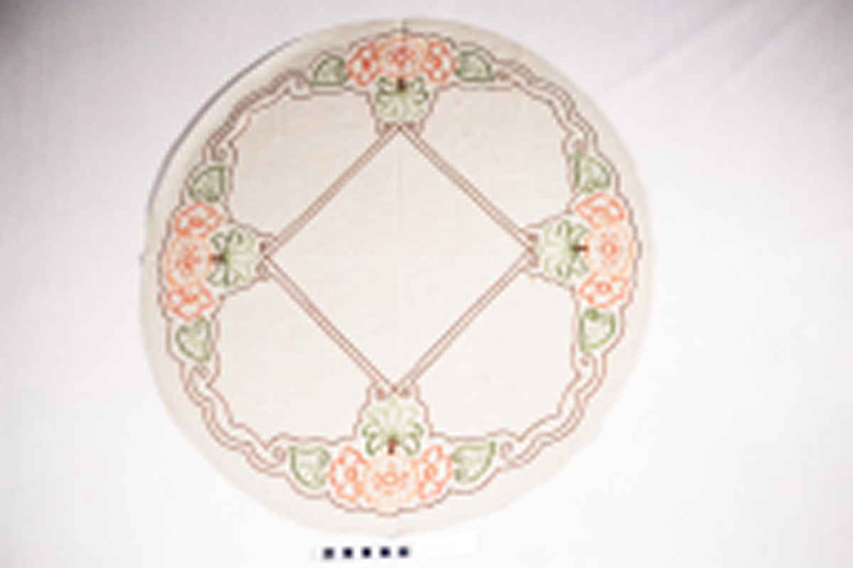 Oval, brodert duk, blomemotiv. Sydd plattsaum og stilkesting.