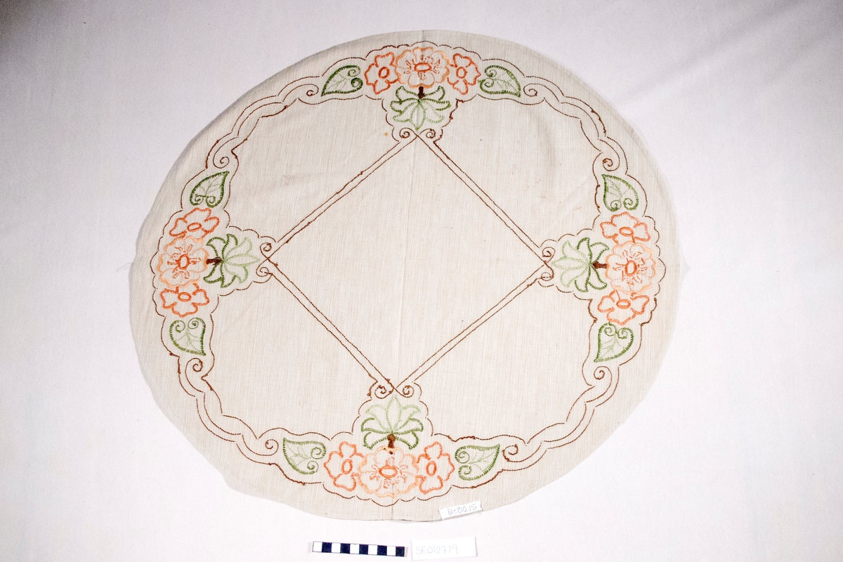 Oval, brodert duk, blomemotiv. Sydd plattsaum og stilkesting.