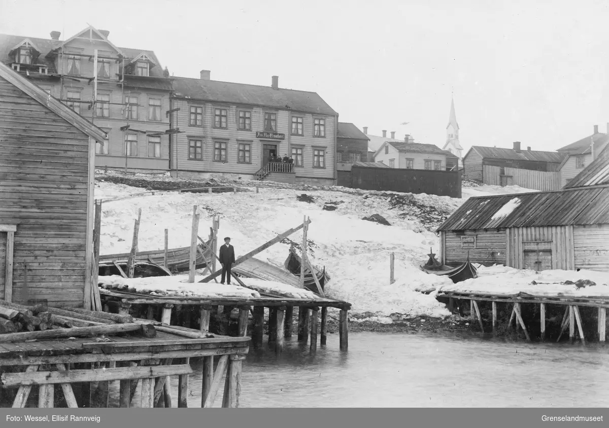 Havnemiljø fra Vadsø. En gruppe mennesker står på trappen til Fru Aas Privat Hotel oppe i bakken. Noen arbeider på taket i nabobygningen. Kirka i bakgrunnen.