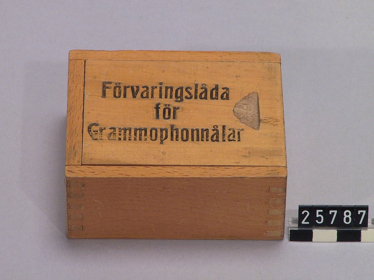 Ask av trä till förvaring av grammofonnålar. Med skjutlock med text: "Ask för Grammophonnålar. Ett fack med halvcylindrisk botten för obegagnade nålar, ett fack med lock med hål för begagnade".