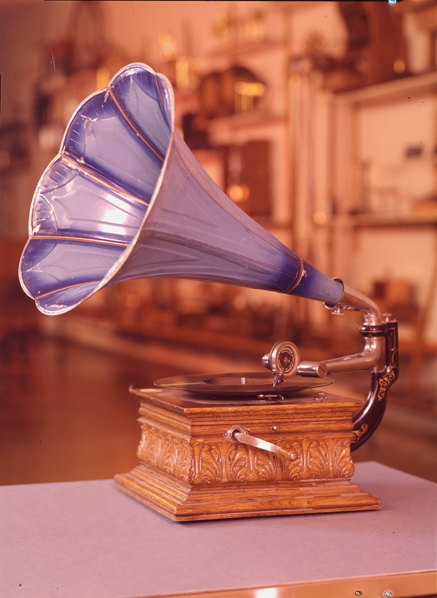 Grammofon med tratt av rödlackerad plåt (tratt TM 34.449). Ljuddosa "Exhibition", tillverkad i USA av Gramophone & Typewriter Ltd (London Berlin Paris). Lådan och troligen verket av Deutsche Gramophon AG. Lådan stämplad i botten "DGAG 7484 Pev K". Skylt med en "skrivande ängel" på lådan. Enkelfjädrigt verk.