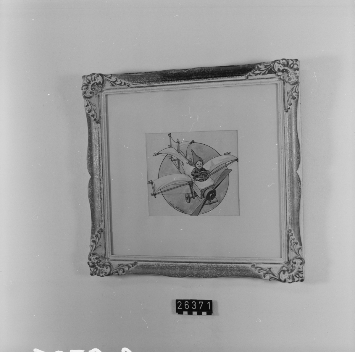 Förlaga till vykort, pojke i tomteluva i improviserad flygmaskin av tidig Bleriot-typ. I ram under glas. Ramen från 1950-talet.