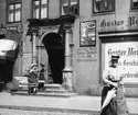 Skioptikonbild med motiv av tre små flickor utanför klädbutik i Lüneburg.
Bilden har förvarats i kartong märkt: Resan 1906. Lüneburg 8.