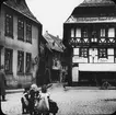 Skioptikonbild med motiv av lekande barn på torg vid Bergstrasse 60 i Goslar. Vid Bahrs Kaffee (kafferosteri).
Bilden har förvarats i kartong märkt: Resan 1907. Gosslar 7.