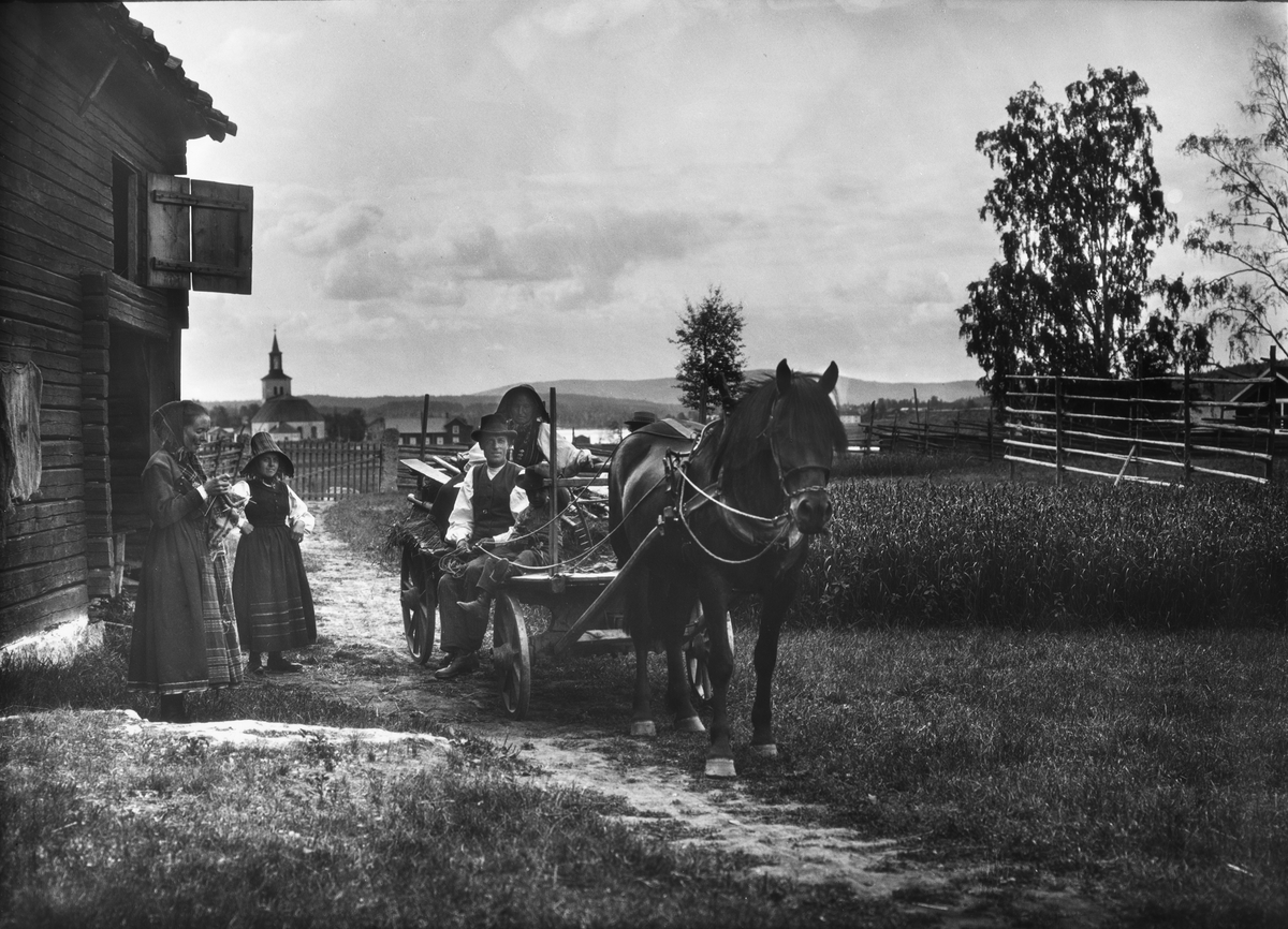 Diapostiv, fönsterbild med motiv från Floda. Tre kvinnot (kullor) i traditionsenlig dräkt, varav en sittande på kärra med man och litet barn, vilken drages av arbetshäst. Kyrka i bakgrunden.