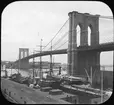 Skioptikonbild med motiv av fartyg i hamn vid Brooklyn Bridge, New York.