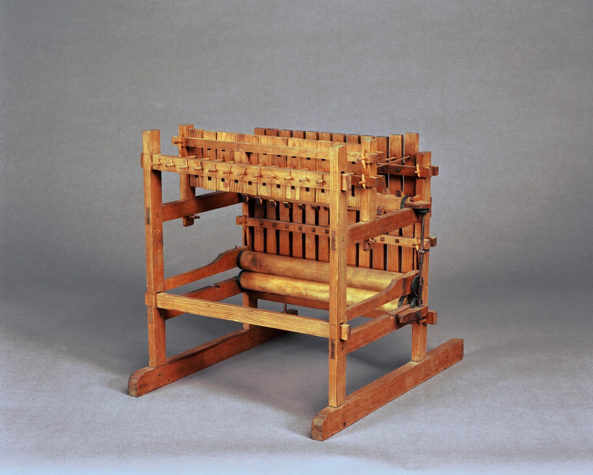 Modell av tröskverk. Text på föremålet: "N:o 52". I Jonas Norbergs förteckning från 1779 beskrivs modellen på följande sätt: " Modell på Trösk Machine med stampar, at drifvas medelst strömfall, gjord af underteknad [J. Norberg], med åtskillige förbättringar. I synnerhet är densamma så inrättad, att om än vatnhjulet ligger aldrig så djupt ned i strömmen, Så står ändå tröskmachinen uppe på jämna marken# och dess kuggevalts som lyfter stamparna, blir medelst en särskilt invention omkringdragen af en, (45 grader eller något mindre) över vatnhorizonten lutande smal stock, som går ner til vatnhjulaxeln såsnart sädeskornen komma lösa utur axen, rinna de straxt ned uti en, under det gående halmlaget belägen, igenlåst brädlår, så at ingen uttröskad säd, kan af otrogit folk bortstjålas. Kunna stångren, på hvilka stamparna hänga, hvar för sig, medelst kilar spännas högre eller lägre, alt som det fordras, på det at stamparne efter slagen icke må hindra det framskridande sädesslaget, och at stångren må lagom trycka stamparna."