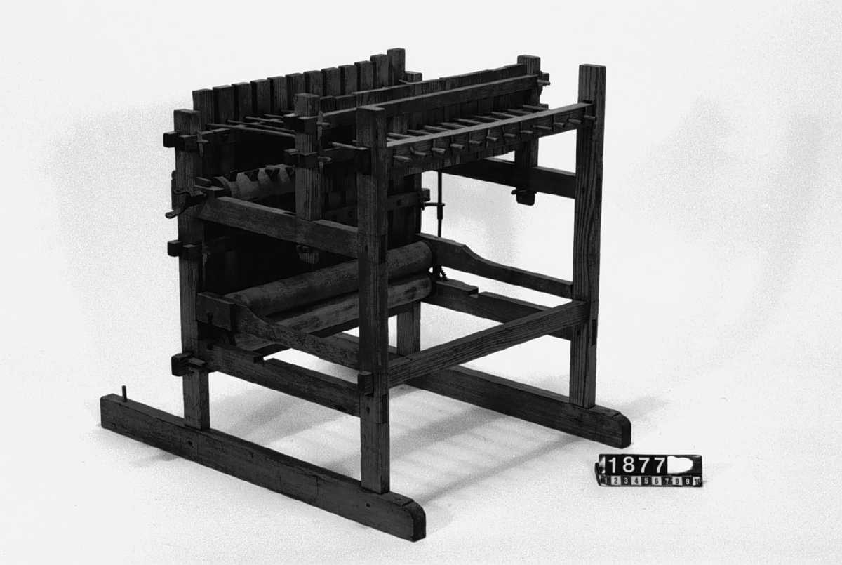 Modell av tröskverk. Text på föremålet: "N:o 52". I Jonas Norbergs förteckning från 1779 beskrivs modellen på följande sätt: " Modell på Trösk Machine med stampar, at drifvas medelst strömfall, gjord af underteknad [J. Norberg], med åtskillige förbättringar. I synnerhet är densamma så inrättad, att om än vatnhjulet ligger aldrig så djupt ned i strömmen, Så står ändå tröskmachinen uppe på jämna marken# och dess kuggevalts som lyfter stamparna, blir medelst en särskilt invention omkringdragen af en, (45 grader eller något mindre) över vatnhorizonten lutande smal stock, som går ner til vatnhjulaxeln såsnart sädeskornen komma lösa utur axen, rinna de straxt ned uti en, under det gående halmlaget belägen, igenlåst brädlår, så at ingen uttröskad säd, kan af otrogit folk bortstjålas. Kunna stångren, på hvilka stamparna hänga, hvar för sig, medelst kilar spännas högre eller lägre, alt som det fordras, på det at stamparne efter slagen icke må hindra det framskridande sädesslaget, och at stångren må lagom trycka stamparna."