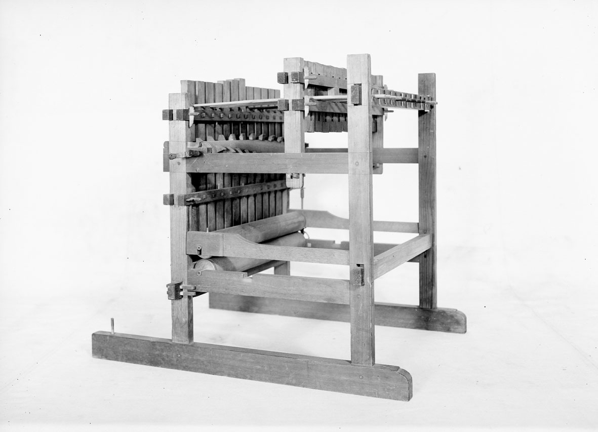 Modell av tröskverk. Text på föremålet: "N:o 52". I Jonas Norbergs förteckning från 1779 beskrivs modellen på följande sätt: " Modell på Trösk Machine med stampar, at drifvas medelst strömfall, gjord af underteknad [J. Norberg], med åtskillige förbättringar. I synnerhet är densamma så inrättad, att om än vatnhjulet ligger aldrig så djupt ned i strömmen, Så står ändå tröskmachinen uppe på jämna marken# och dess kuggevalts som lyfter stamparna, blir medelst en särskilt invention omkringdragen af en, (45 grader eller något mindre) över vatnhorizonten lutande smal stock, som går ner til vatnhjulaxeln såsnart sädeskornen komma lösa utur axen, rinna de straxt ned uti en, under det gående halmlaget belägen, igenlåst brädlår, så at ingen uttröskad säd, kan af otrogit folk bortstjålas. Kunna stångren, på hvilka stamparna hänga, hvar för sig, medelst kilar spännas högre eller lägre, alt som det fordras, på det at stamparne efter slagen icke må hindra det framskridande sädesslaget, och at stångren må lagom trycka stamparna."