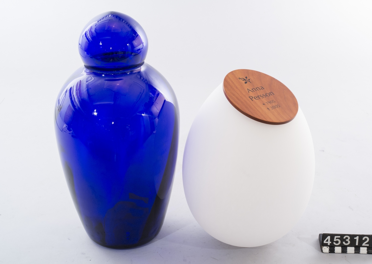 Nedbrytningsbar askurna av patenterat glasmaterial som vittrar sönder efter gravsättning. Då urnan med aska utsätts för markens fukt startar en process som bryter ner glasets grundstruktur. Efter 15 år har glaset omvandlats till grus.  Två urnor - en blå med lock av glas modell Andante samt en matt vit modell Dolce med lock av trä märkt "Anna Persson *1905 +2005", B-37.  Båda märkta "Mats Jonasson, Målerås".