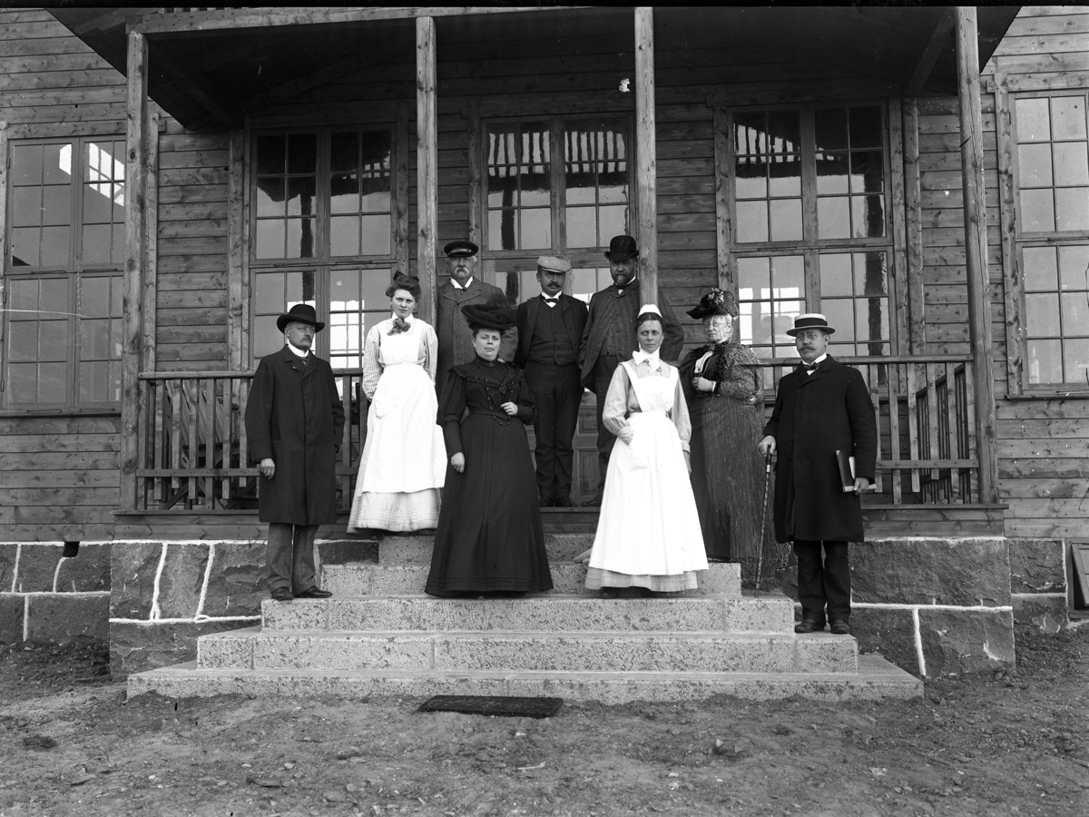 En grupp personer på trappan till en veranda på Kustsanatoriet Apelviken år 1907. Personal och troligen personer i styrelsen för "Föreningen för kustvård åt skrofulösa barn" som drev Kustsanatoriet. Den första vårdpaviljongen uppfördes 1904 men fler krävdes i rask takt. Att döma av grundfogarna, trappan och marken runtom är denna byggnad nyligen uppförd, så kanske är bilden tagen i samband med en invigning av densamma.