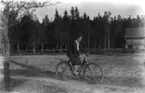 En flicka på cykel. Bostadshus till höger på bilden.
Thea Elfrida Olsson, Erik Theon Olssons dotter.