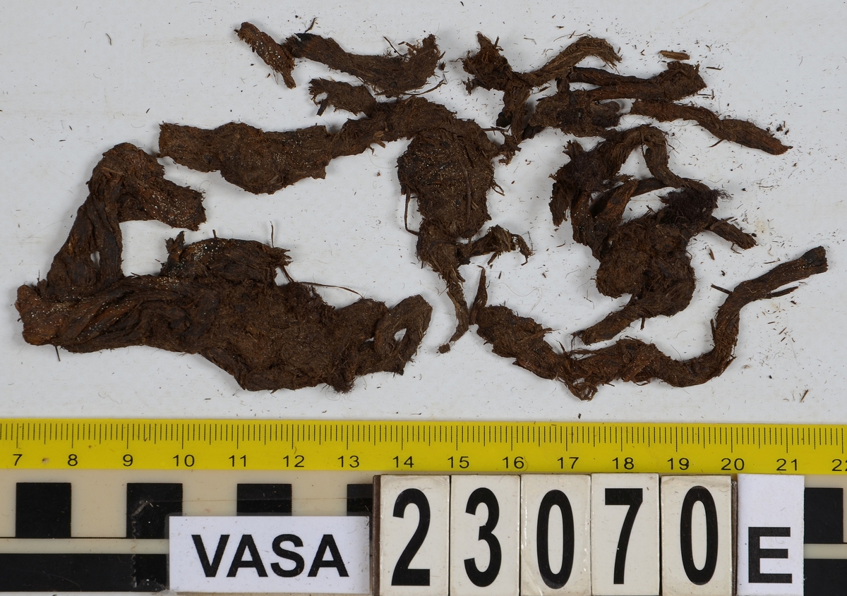 Sko.
Inuti skorna hittades även fragment från olika textilier som ursprungligen varit ett par strumpor. Idag sitter ett stickat fragment av ull fast i vänsterskon (23070) med avigsidan uppåt. I fyndet finns även en mängd lösa stickade fragment som idag är mycket korroderade och vissa helt mineraliserade av järn vilket har gjort textilen stenhård och därmed oerhört skör (23070a-c). Utifrån minskningar som några små fragment har så har det varit möjligt att rekonstruera strumpans tå. Genom att pussla ihop fragmenten som ursprungligen har suttit ihop har det på så vis gått att avgöra hur strumpans tå har varit konstruerad. Då textilen är så fragmentarisk är det inte möjligt att avgöra hur resten av strumpan har sett ut eller varit konstruerad. Däremot så finns det fragment av ett kypertvävt tyg av ull som på vissa ställen ligger dubbelt med det stickade 23070c). Ett av dessa fragment har stelnat i formen så som den suttit på fotens främre del inuti skon. Detta fragment täcker delvis det stickade tyget och har dessutom spår av kaststygn längs en kant. Så som det vävda tyget är placerat på det stickade tyget pekar på att man använt det som lagningslappar till sina stickade strumpor. 
I fyndet finns det även korroderade järnklumpar, ett tuskaftsvävt tyg av ull och rester av ett grovt bastfibergarn och två mindre läderfragment (23070d-f). Det är möjligt att även dessa har tillhört strumpan och/eller skon.

För mer detaljerad information från textildokumentationen (år 2022) se länkade filer.

Skodokumentation 2022:
Ovanläder. Tillverkad i getskinn av god kvalitet. Hör ihop med fyndnummer 23069 (Yngves skor). Tillverkades på en rak läst och bars på vänster fot. Inga dekorationer Det finns stygnhål som visar att det funnits invändiga förstärkningar (men förstärkningslappen saknas). Det finns delaminering men inga lagningar.
För mer information, se Länkade filer: Uppritning

Text in english:
This is the upper of a shoe of more “extreme fashion”, made of goatskin leather of good quality, probably made on a straight last and worn of the left foot, with no decorative features. The upper couples with 23069, together composing Yngve’s shoes. There are internal reinforcements missing from the upper and delamination, there are no repairs.
For more information, go to Länkade filer: Uppritning