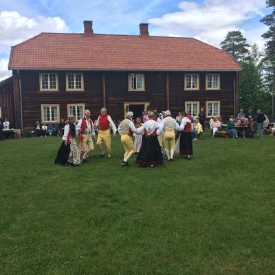 Folkedans på tunet under Liv i stuene. I bakgrunnen ser vi staselige Gjersøyen, et tømmerhus med to etasjer.