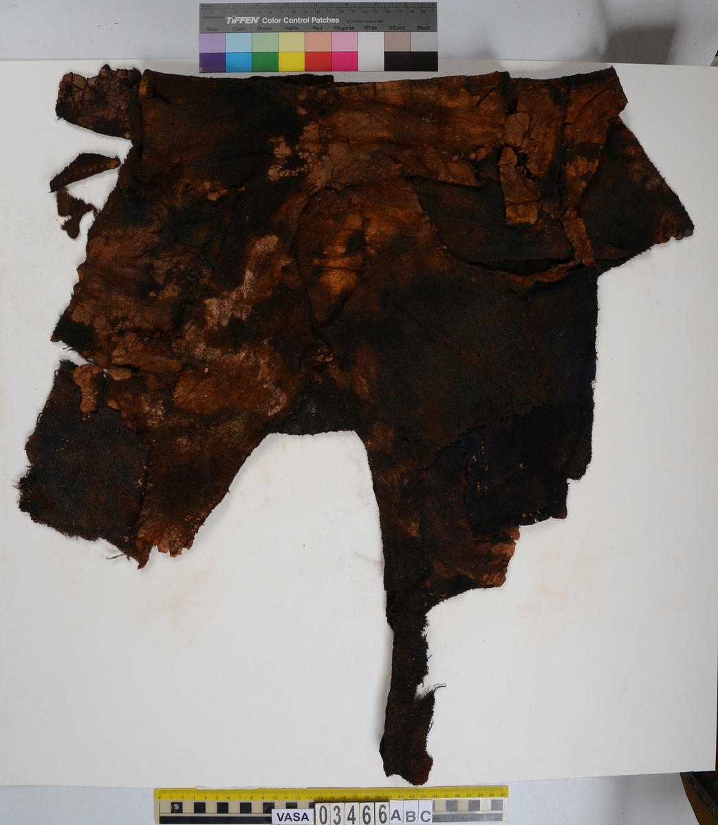 Textil. 13 tygfragment uppdelade på fyndnummer 03466a-d.
Fnr 03466a består av 10 fragment av ull vävda i tuskaft varav det största är ihopsytt av flera delar. Har varit ett par byxor med lucka fram. Originalfärgen kan ha varit blå då rester av blå färg syns på insidan av två av bitarna på de delar där sömmen varit och tyget originalyta bevarats bättre. På en av dessa bitar som tolkats som en del av en ficka på byxans vänstra sida finns rester av en tvåtrådig, s-tvinnad, z-spunnen tråd i sömmen. Byxans tyg har varit mer valkad på insidan än på utsidan. På det största fragmentet framsida syns två knapphål i linningen, och rester efter ytterligare två hål. Det kan därför konstateras att det ursprungligen har varit två knapphål på var sida på linningens mittdel där sidodelarna av linningen knäppts eller knutits fast. Vid ena knapphålet syns något som kan vara resterna av en knapp eller hyska odyl. När byxans delar läggs ihop som de en gång suttit mäter midjebredden ca 47 cm vilket ger ett midjeomfång på ca 94 cm före ihopsnörning. Av det som kvarstår av byxbenen kan konstateras att dagens (år 2015) längd på vänster byxben är 60 cm från linningens övre kant och ner. Höger byxben är i dagsläget endast 43 cm långt. 
På fnr 03466b syns rester av söm i en av kanterna. Fragmentet är av ull och vävt i 2/2-kypert. Lappen har en söm i en cirkel mitt på tyget som troligen är resterna efter den söm som sytt fast lappen runt ett hål på byxan. Kan ha varit en lagningslapp på byxans högra knä som uppvisar motsvarande söm kring ett cirkelformat hål. 
Fnr 03466c är ett fragment av ull vävt i lite finare tuskaftsväv än fnr 03466a. Fragmentet har hål efter sömmar i kanterna samt delar av en ljus tråd i en av kanterna. Blå färg syns där tyget varit skyddat där sömmen varit. Fragmentet kan ha varit en innerficka bak på byxans vänstra sida. Det finns en söm i byxans tyg där som överensstämmer med lappens söm.
Alla kanterna på fnr 03466d är invikta och har hål efter söm som om biten varit fastsydd på ett annat tygstycke. Fragmentet är av ull och vävt i samma sorts tuskaftsväv som byxan. Kan ha varit en lagningslapp på byxan över ett hål i byxtyget på den plats där vänster innerficka bak är placerad.
